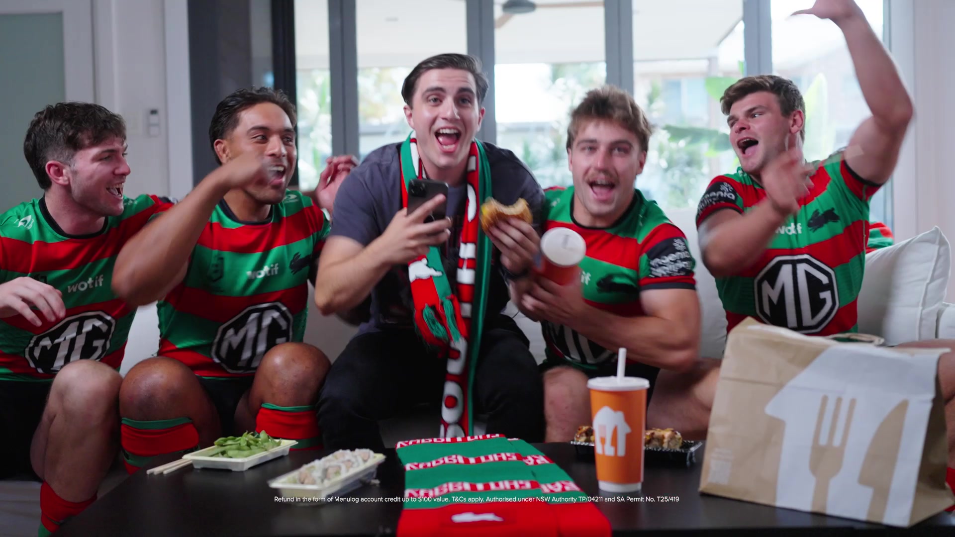 16:9 frame: all five on the couch celebrating, the Menulog bag and drink on the table