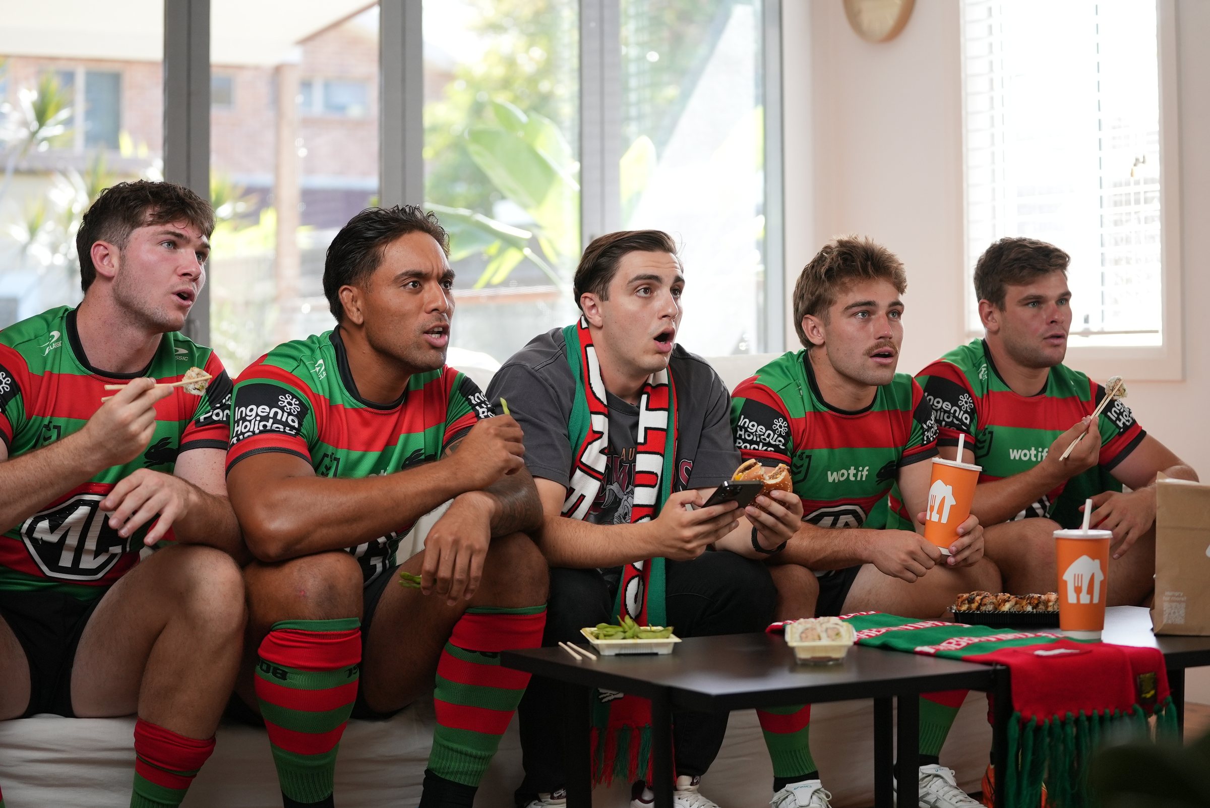 The fan in a Rabbitohs scarf on the couch with four Rabbitohs players, eyes on the game