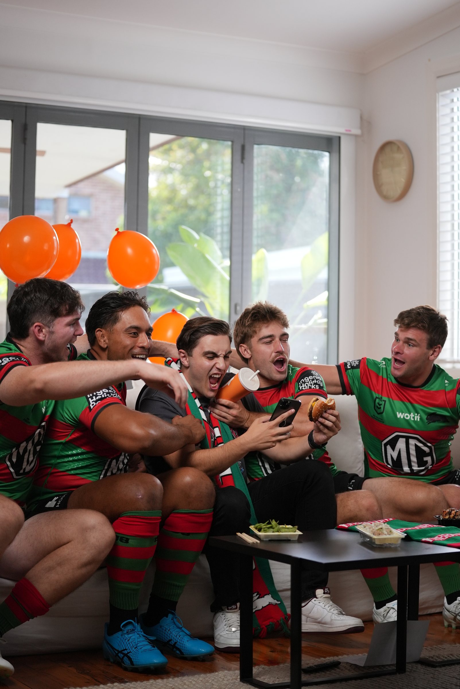 The try goes in: the players pile onto the fan, who holds up his phone, burger and Menulog cup, orange balloons behind