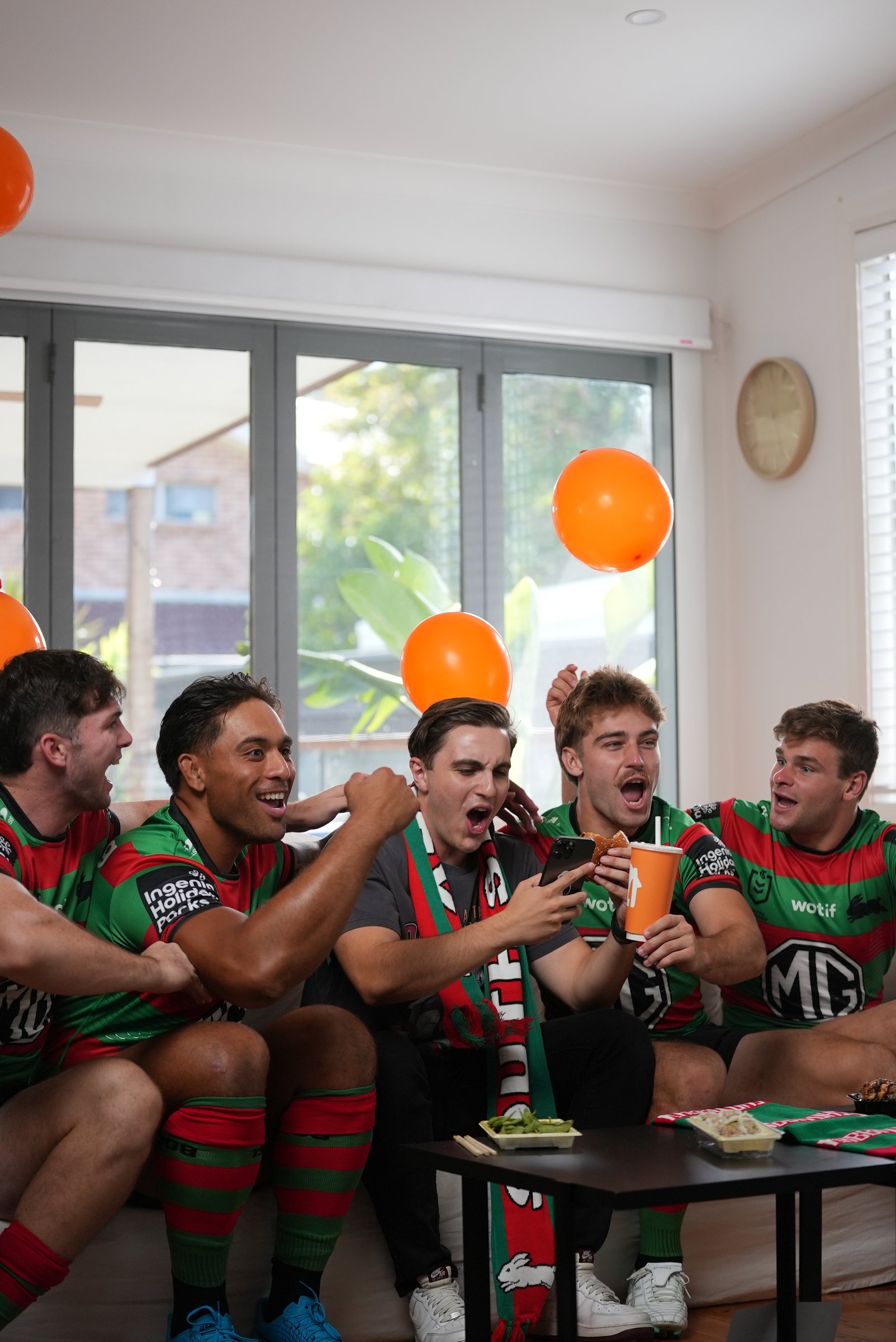 The players celebrating around the fan as he checks his phone, Menulog cup in hand, balloons in the air